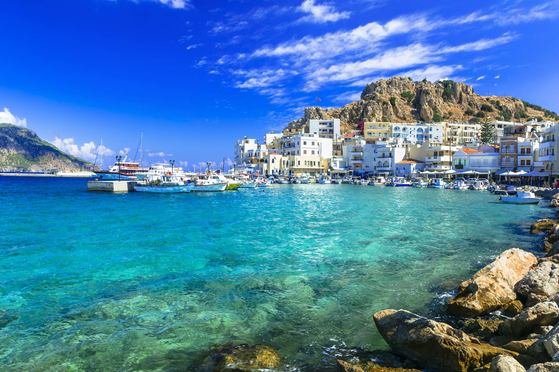 Turkusowa woda, zacumowane łodzie i białe budynki na wzgórzu – tak wygląda port w Pigadia na Karpathos.