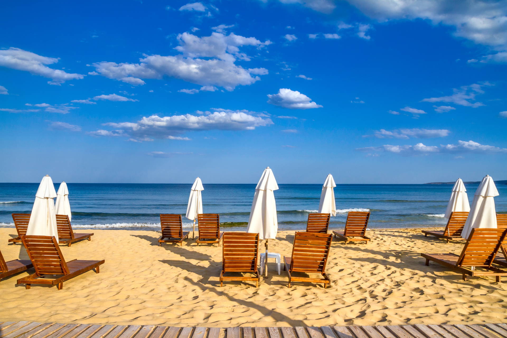 Drewniane leżaki i białe parasole na piaszczystej plaży nad morzem w słoneczny dzień.
