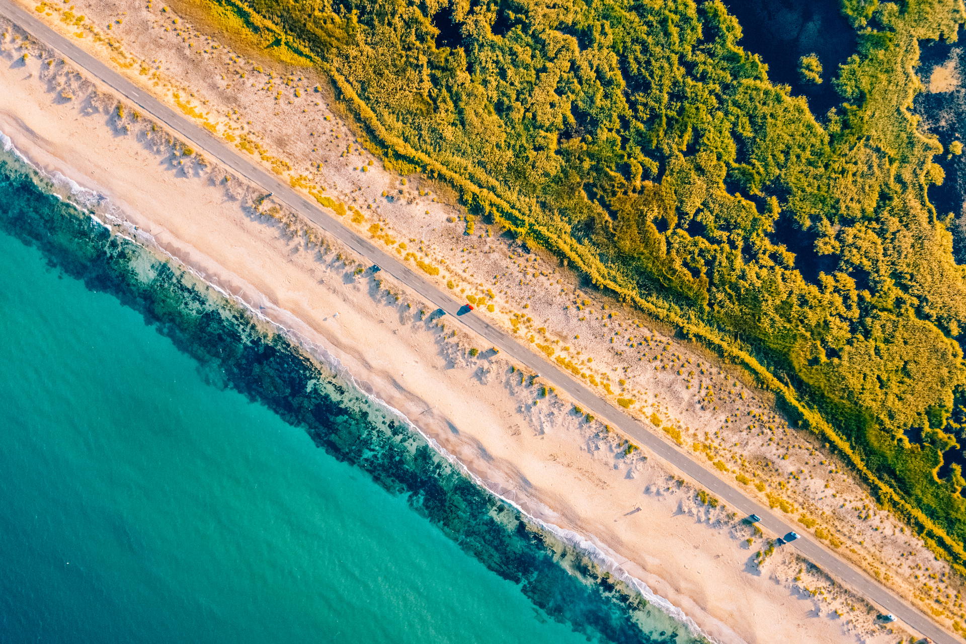 Z lotu ptaka widać wybrzeże: turkusowe morze, piaszczystą plażę, drogę i zielone mokradła.
