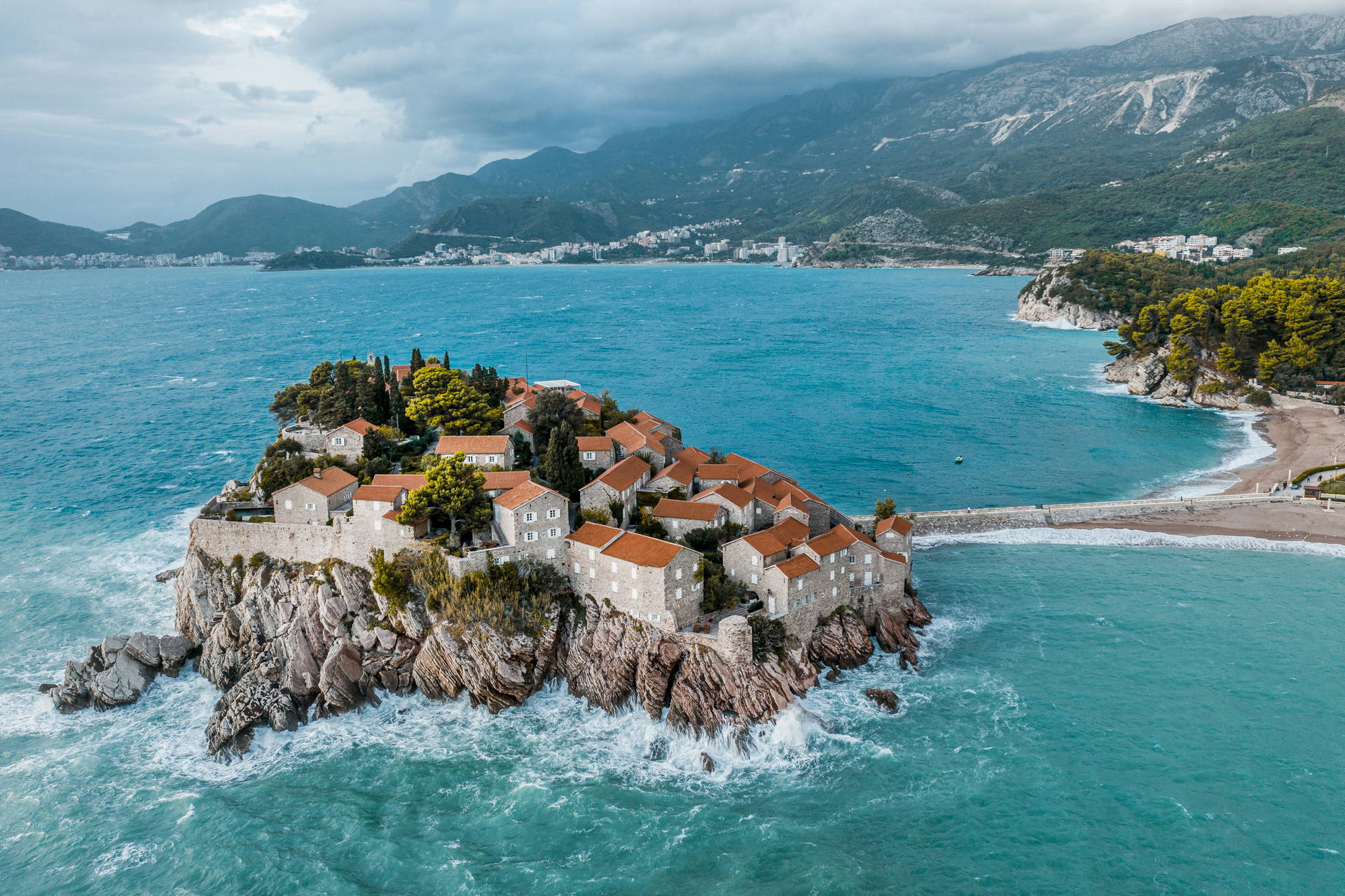 Widok z lotu ptaka na wyspę Sveti Stefan w Czarnogórze, połączoną z lądem groblą i zabudowaną kamiennymi domami z czerwonymi dachami.