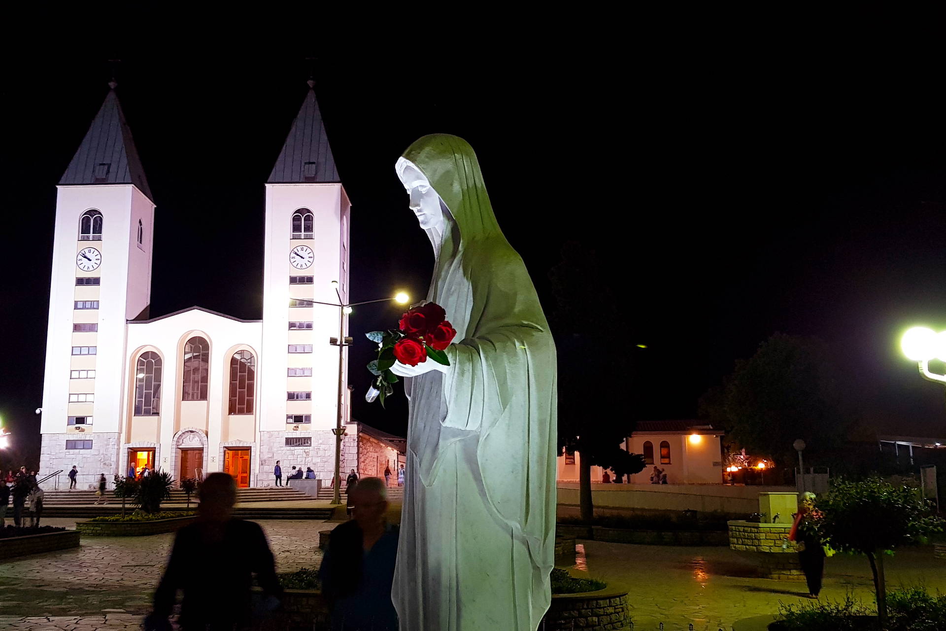 Figura Matki Bożej z różami na tle kościoła św. Jakuba w Medziugorie nocą.