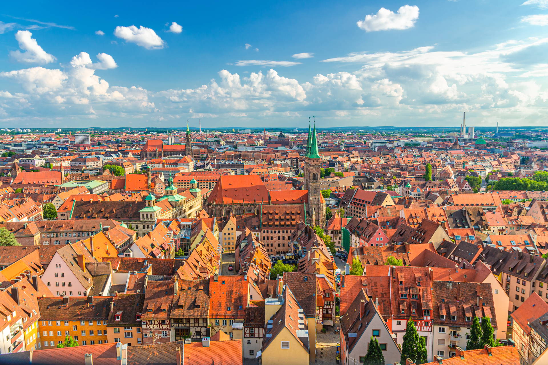 Widok z góry na stare miasto w Norymberdze. Gęsta zabudowa z czerwonymi dachami i wieżami kościołów na tle błękitnego nieba.