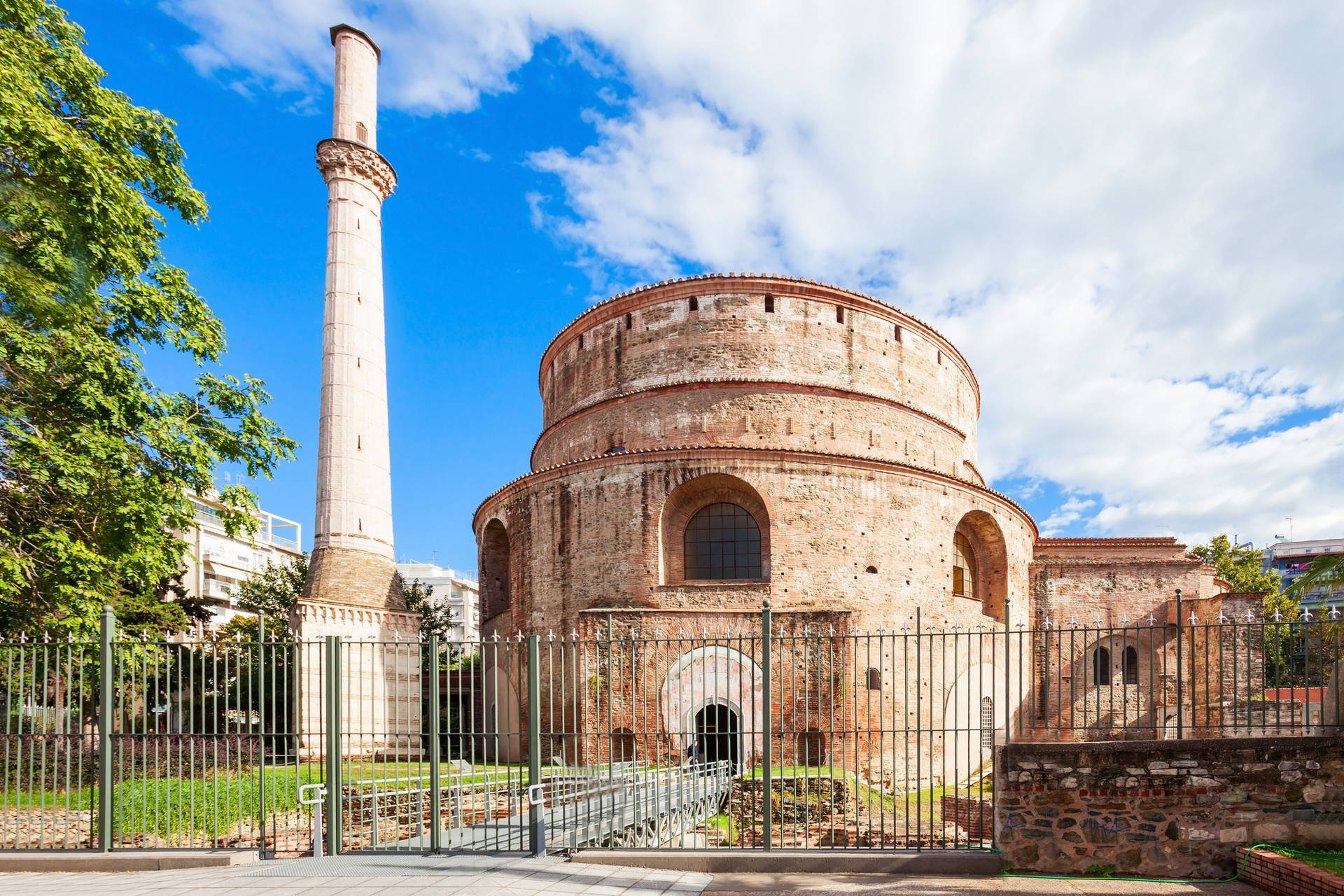 Rotunda w Salonikach, ceglany budynek na planie koła z wieżą, otoczony metalowym ogrodzeniem.