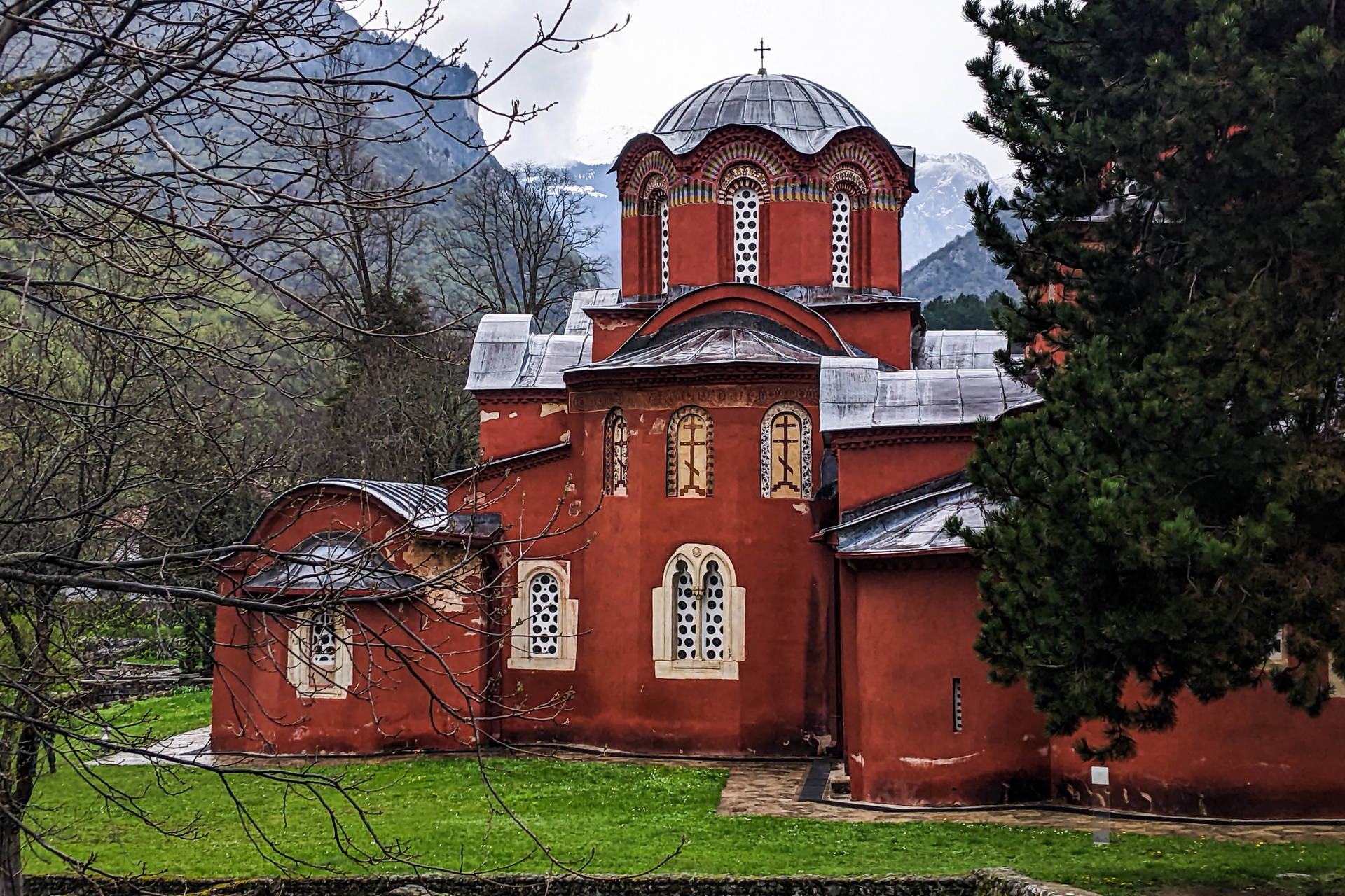 Czerwony budynek z kopułą w Monasterze Peć w Kosowie, na tle zieleni i ośnieżonych szczytów gór.