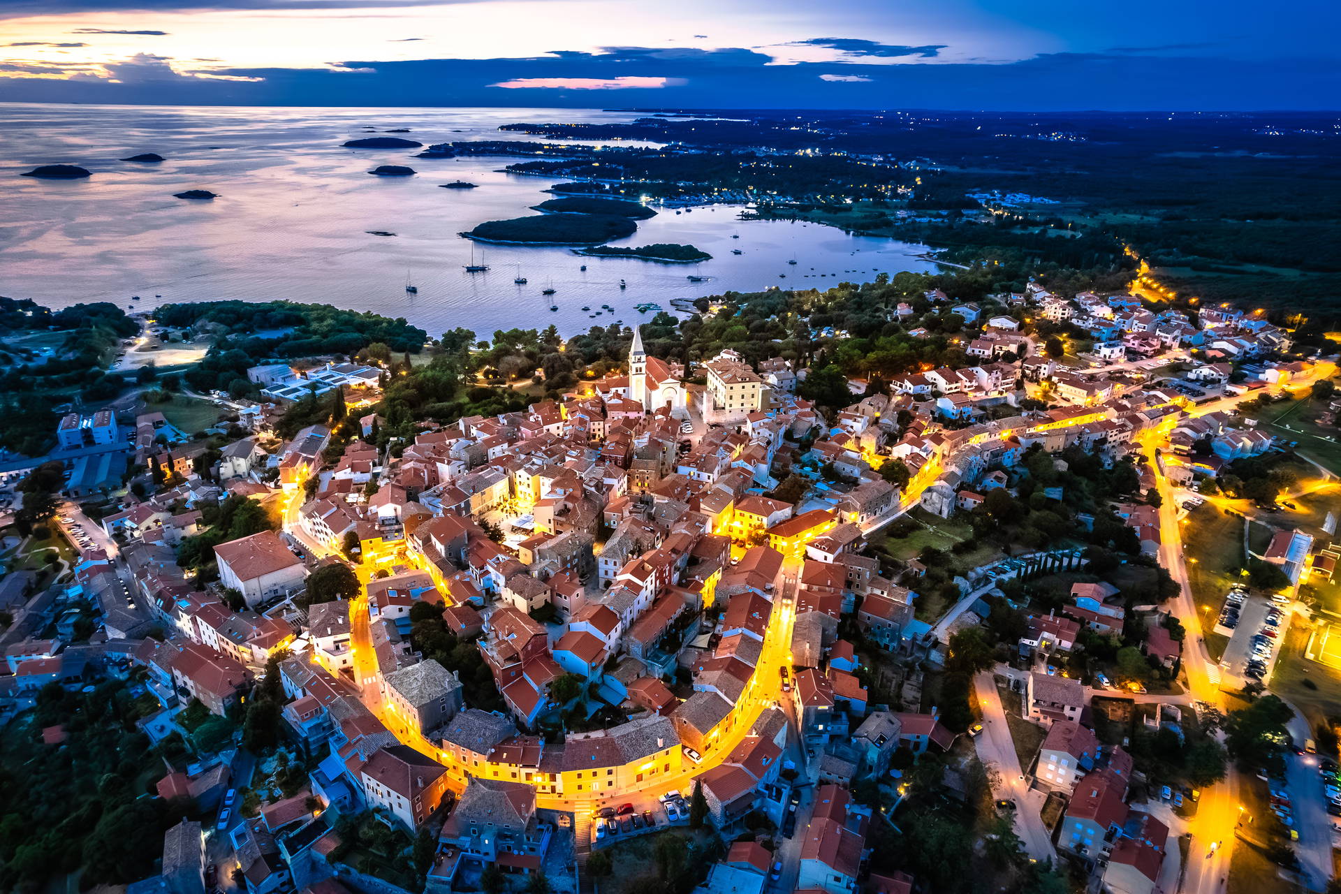 Widok z lotu ptaka na oświetlone miasto Vrsar w Chorwacji o zmierzchu. W tle widać morze i archipelag.