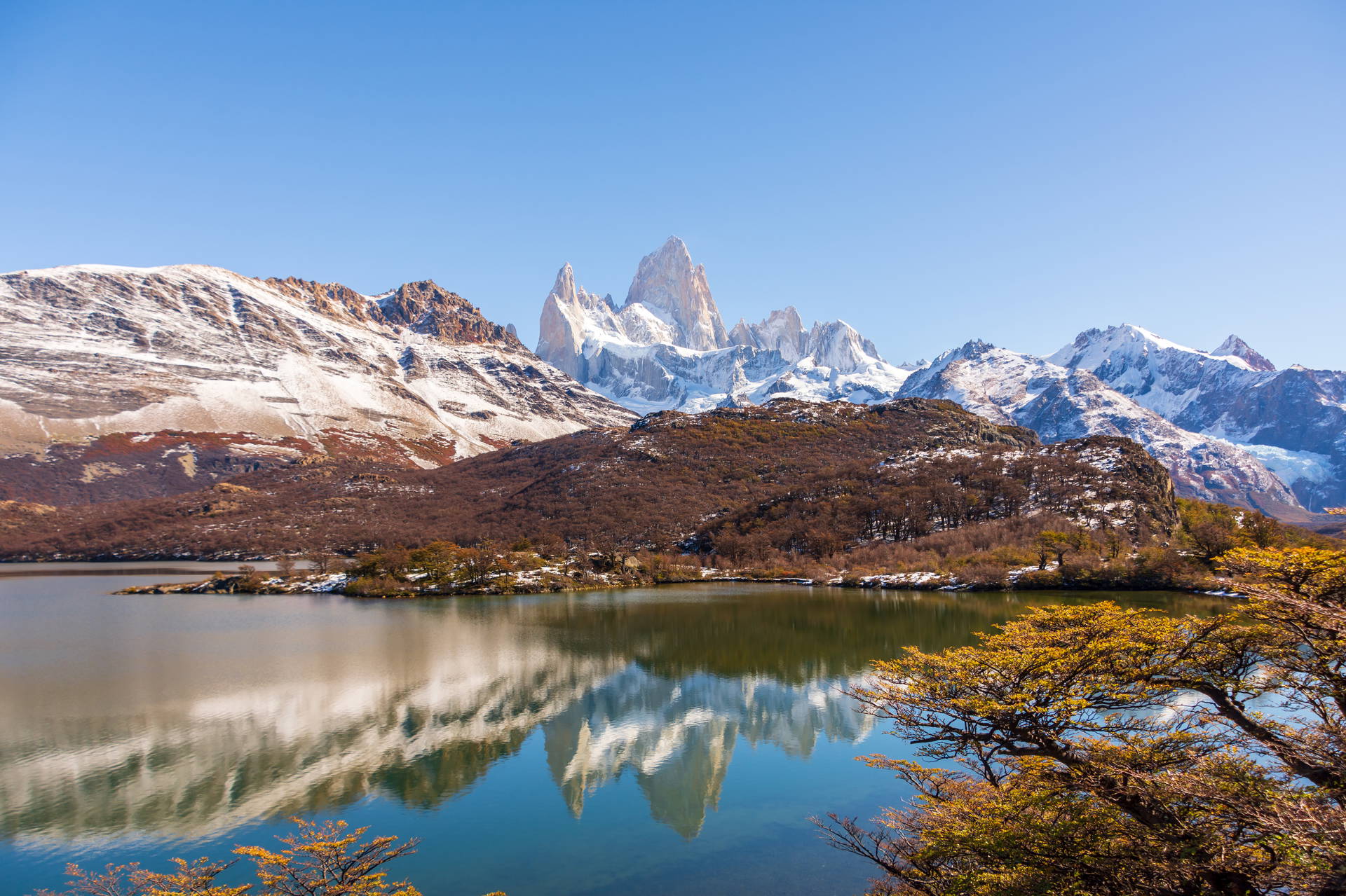 Góra Fitz Roy w Argentynie odbijająca się w jeziorze, otoczona ośnieżonymi szczytami i jesiennym lasem.