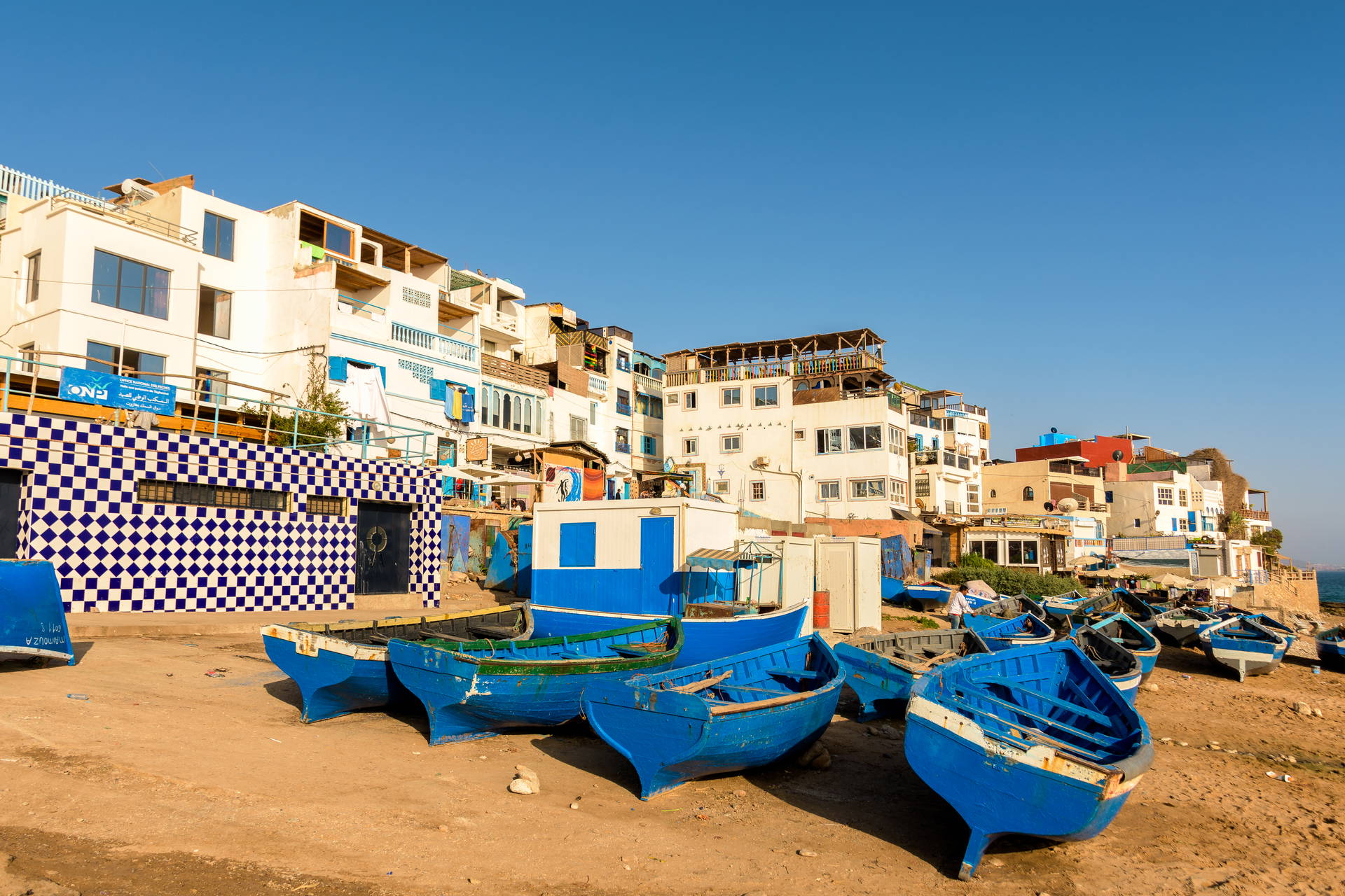 Niebieskie łodzie rybackie na piaszczystej plaży w Taghazout w Maroku. W tle białe budynki miasteczka.