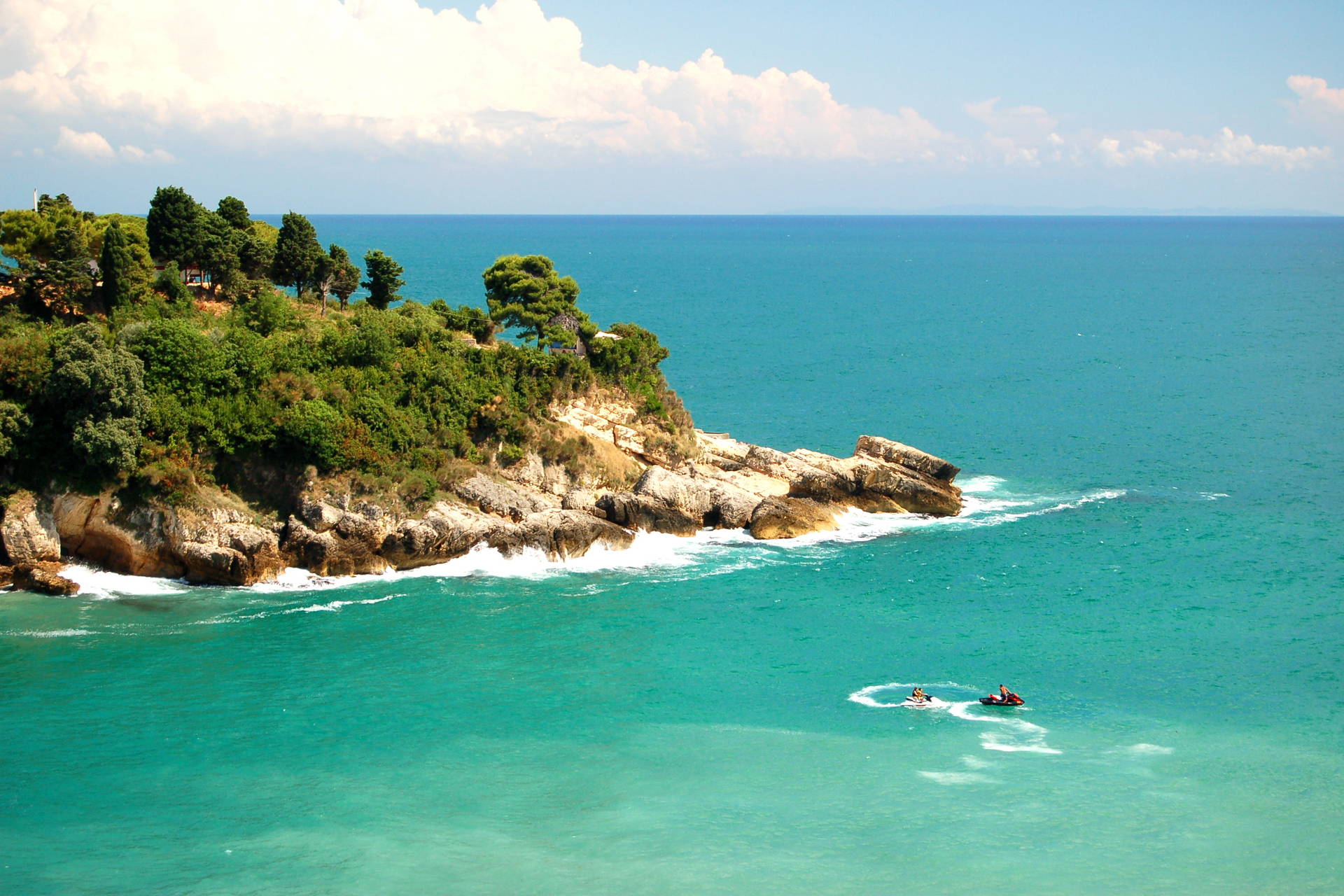 Skalisty klif porośnięty zielenią nad turkusowym morzem, po którym pływają dwa skutery wodne.