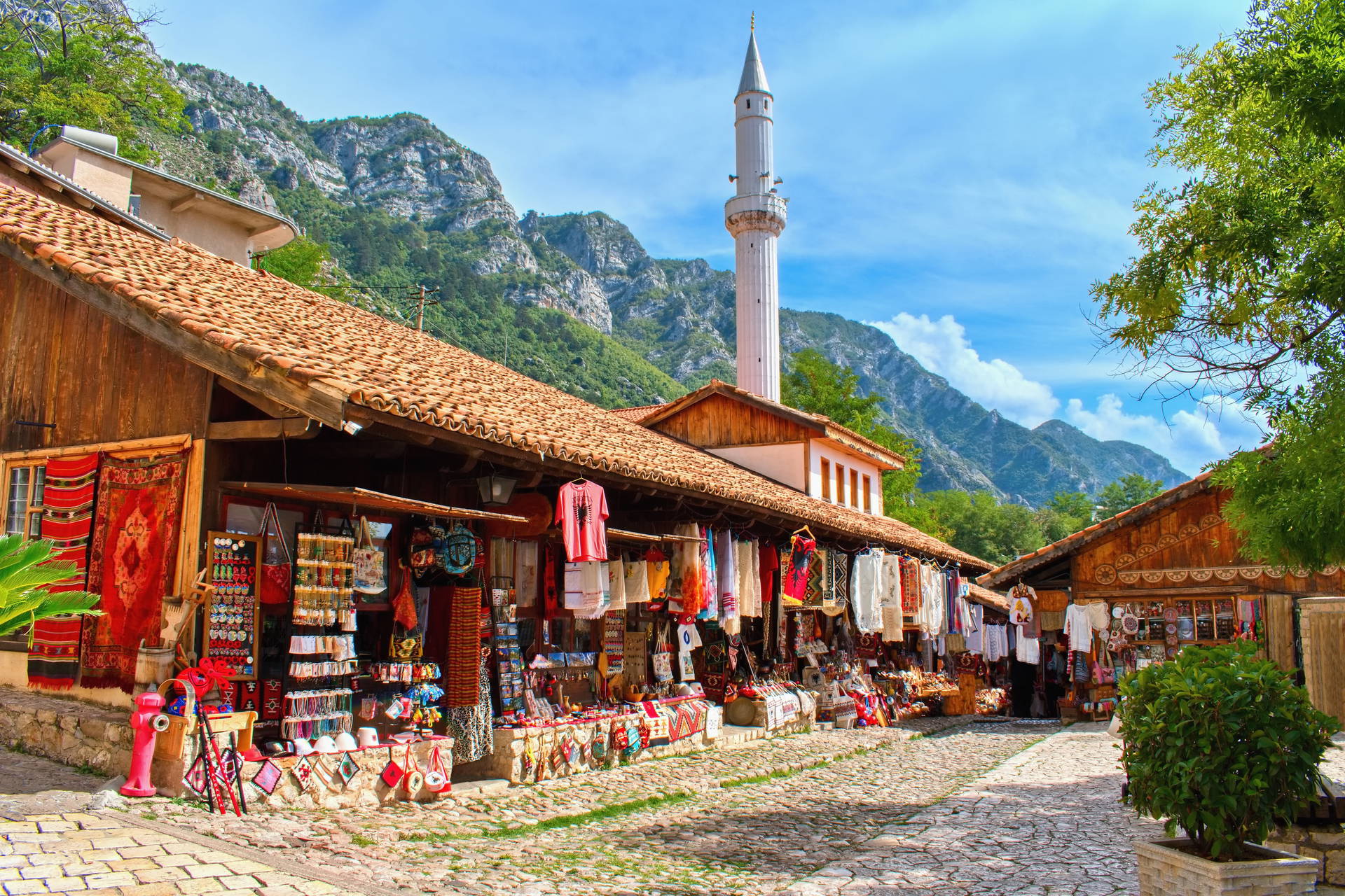 Stary Bazar w Krui w Albanii. Brukowana uliczka z kramami z pamiątkami, a w tle góry i minaret meczetu.
