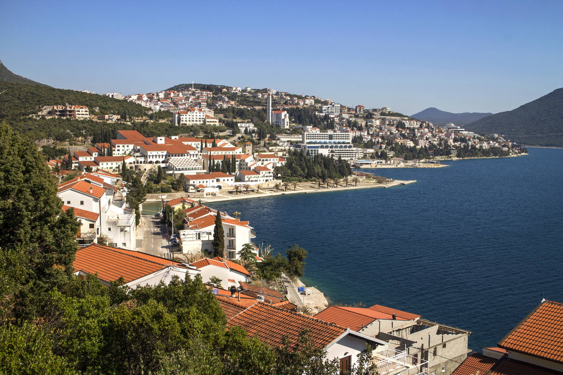 Panoramiczny widok na Neum – miasteczko z białymi budynkami i czerwonymi dachami, położone na wzgórzu nad błękitnym morzem.