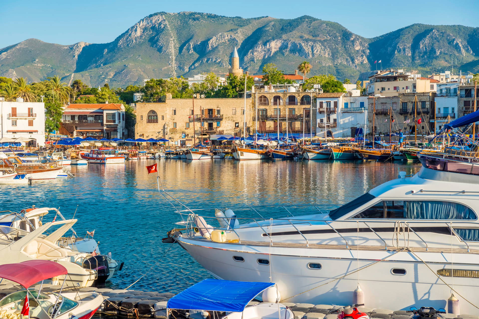 Port w Kyrenii pełen łodzi i jachtów. Wzdłuż nabrzeża stoją kamienne budynki, a w tle widać góry.