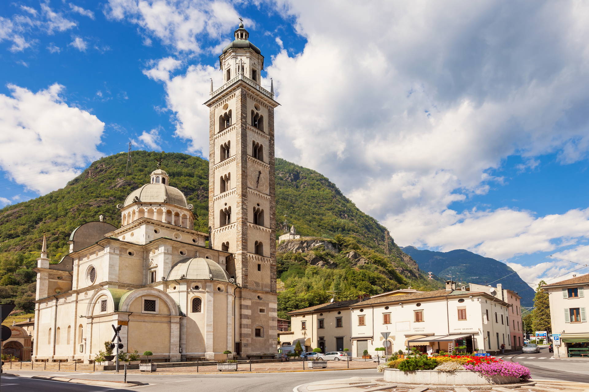 Sanktuarium Madonna di Tirano z wysoką dzwonnicą na tle zielonych gór w słoneczny dzień.