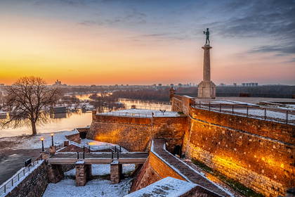 Pomnik Zwycięzcy w twierdzy Kalemegdan w Belgradzie o zimowym wschodzie słońca. W tle widać ośnieżone mury i rzekę.