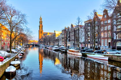 Ośnieżony kanał w Amsterdamie w zimowy poranek. W tle widać wieżę kościoła Westerkerk i jej odbicie w wodzie.