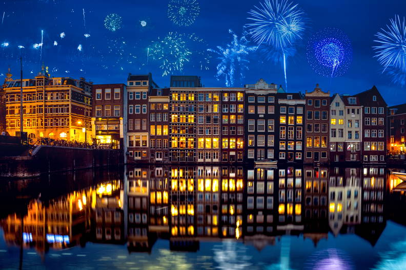 Nocny pokaz fajerwerków nad kamienicami w Amsterdamie, którego światła odbijają się w kanale.