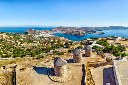 Trzy kamienne wiatraki na wzgórzu greckiej wyspy Patmos, z widokiem na zatokę i miasto.