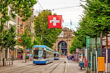 Ulica w Zurychu z niebieskim tramwajem i flagą Szwajcarii. W tle widać dworzec Bahnhof Enge.