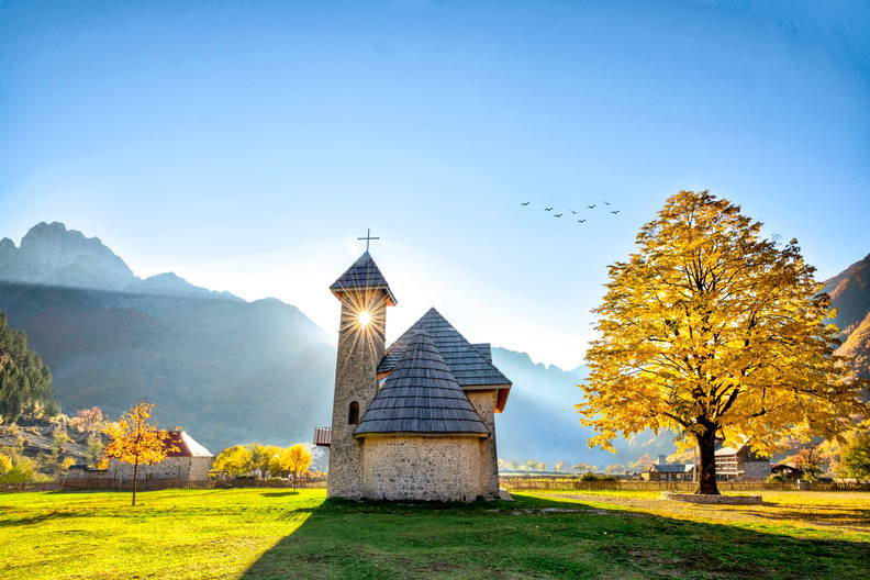 Kamienna cerkiew w Theth na tle gór i drzewa z żółtymi liśćmi.