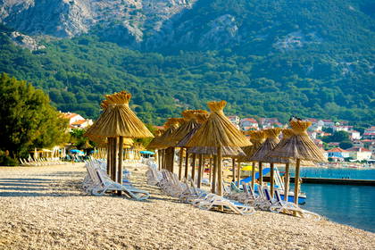 Kamienista plaża w Chorwacji z rzędami słomianych parasoli i leżaków na tle gór.