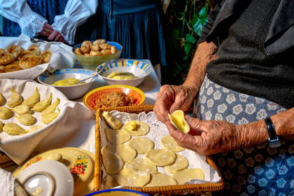 Starsza osoba lepi pierogi przy stole zastawionym jedzeniem.