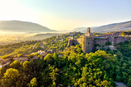 Zamek Gjirokastra z wieżą zegarową na wzgórzu o wschodzie słońca. Poniżej miasto położone w dolinie, w tle góry.
