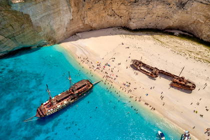 Zatoka Wraku na Zakynthos widziana z lotu ptaka. Na plaży leży zardzewiały statek, a w lazurowej wodzie pływają turyści.
