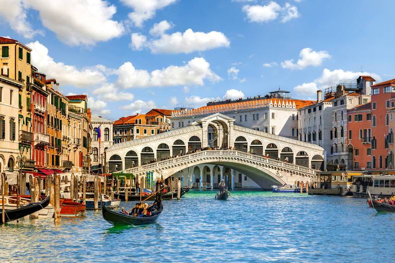 Most Rialto nad Canal Grande w Wenecji w słoneczny dzień. Po kanale pływają gondole, a wzdłuż brzegów stoją kolorowe kamienice.