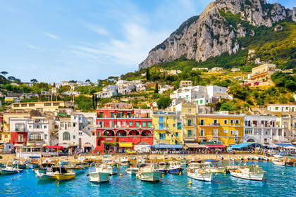 Port Marina Grande na Capri z kolorowymi domami, łodziami na błękitnej wodzie i górą w tle.