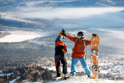 Narciarz i snowboardzista przybijają sobie piątkę na szczycie góry, w tle zimowy krajobraz.