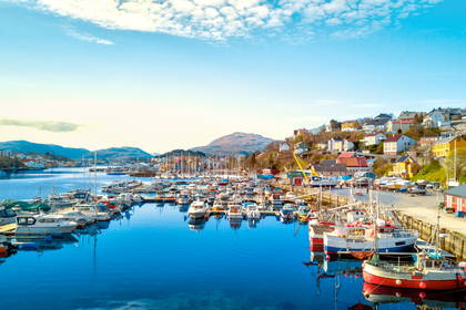 Słoneczny port w Kristiansund w Norwegii. Wiele łodzi i jachtów cumuje w błękitnej wodzie u podnóża wzgórza.