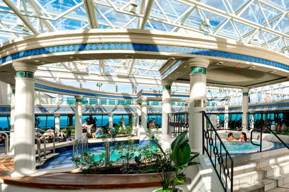 Solarium na statku wycieczkowym pod szklanym dachem, gdzie pasażerowie odpoczywają przy basenach i jacuzzi.